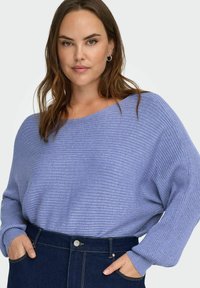 Helelilla soonikkoesemega kampsun laia kaelusega ja pikkade varrukatega, kombineeritud tumesiniste kõrge vöökohaga teksapükstega, millel on esimesed taskud.