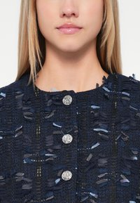 Navy zila zīda žakete ar teksturētu audumu un saplēstiem akcentiem. Iekļauj sudraba pogas un apaļu kakla izgriezumu, radot strukturētu siluetu.