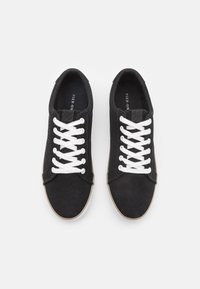 Zapatillas de lona negras con cordones blancos, que presentan un diseño de perfil bajo, puntera redondeada y suela de goma marrón. El interior tiene una plantilla con marca.