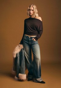 Maglietta oversize marrone con spalle scoperte, jeans a gamba larga blu scoloriti, mentre tiene in mano una giacca di jeans con polsini foderati in pelliccia. Sfondo marrone.