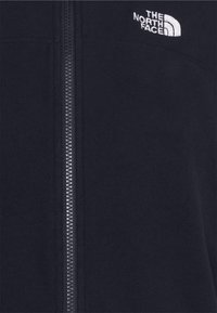 Mörk marinblå fleecejacka med en central dragkedja och vit "The North Face"-logotyp broderad på övre vänstra bröstet.