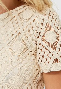 Beige virkattu toppi, jossa on monimutkaisia geometrisia kuvioita auringonsäteillä, avonaiset yksityiskohdat ja lyhyet hihat, jotka luovat tekstuuritun ulkonäön.