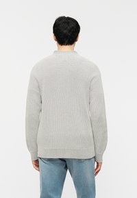 Άνδρας που φοράει ανοιχτόγκρι πλεκτή μπλούζα και μπλε τζιν, στέκεται με την πλάτη στραμμένη σε ένα απλό λευκό φόντο.