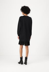 Sweatshirt noir surdimensionné avec des manches longues, des poignets côtelés et une jupe plissée noire en dessous, porté avec des bottines noires à la cheville.