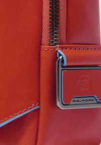 Borsa rossa in pelle con una superficie liscia, dotata di una cerniera nera e dettagli blu. Logo metallico attaccato vicino al cursore della cerniera.