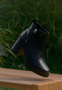 Bottine noire en cuir lisse, dotée d'une fermeture éclair latérale, d'un talon épais empilé et d'un bout arrondi. Présentée sur un fond naturel.