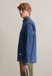 Donkerblauw denim overhemd met een kraag, lange mouwen en knoopdetails aan de manchetten, gecombineerd met olijfgroene broek.
