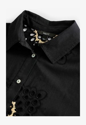 Czarna koszula z ostrym kołnierzykiem, ozdobiona haftowanymi wzorami kwiatowymi i wycięciami. Guziki są jasnego koloru. Materiał wydaje się teksturowany.