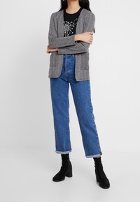 Šedý herringbone blazer s otevřeným přední dílem a náprsními kapsami, přes černé tričko s grafikou, spojený s světle modrými kalhotami s vysokým pasem a černými podpatky.