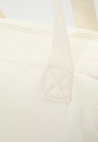 Sac en tissu blanc avec une surface texturée. Dispose d'une fermeture éclair sur le dessus et de poignées plates en tissu. Couture visible près de l'attache des poignées.