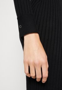 Mano appoggiata di lato, indossando un maglione a maniche lunghe nero a costine con un piccolo logo in metallo sul polsino.