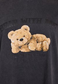 Dark grey t-shirt featuring a graphic of a light brown teddy bear with a smiling expression and text that reads “BEAR WITH ME” above it.