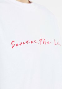 White cotton t-shirt featuring red cursive text “Senses. The Look.” Positioned centrally, smooth texture with a classic crew neck design.