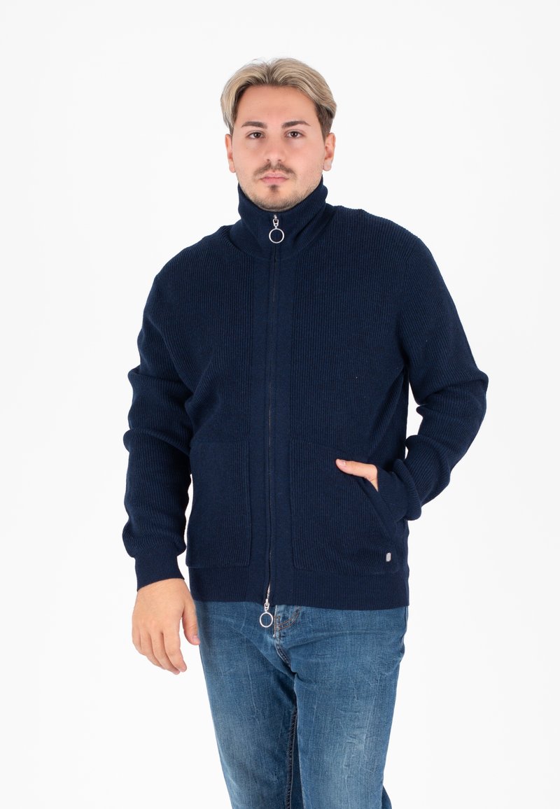 Maglione zip-up blu navy con colletto alto, texture a costine, due tasche frontali e dettaglio della cerniera in argento. Indossato sopra dei jeans blu.