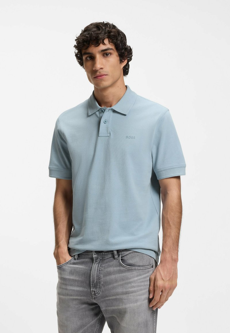 Jeune homme aux cheveux bouclés foncés portant un polo bleu clair à manches courtes et un jean gris, debout avec une main dans la poche.