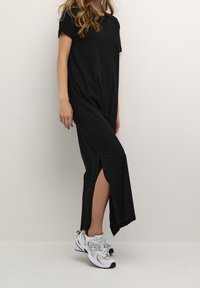 Robe noire longueur genou avec une coupe décontractée et des manches courtes, présentant une fente latérale. Associée à des baskets de sport argentées et noires.