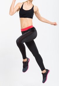 Czarny stanik sportowy z różowym pasem, dopasowane czarne legginsy z boczną siateczką oraz czarne buty sportowe z różowymi podeszwami, w pozie w trakcie skoku.
