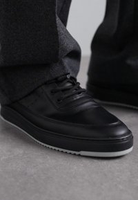 Svart lädersneaker med en slät yta, rund tå och vit gummisula. Har snören och subtila perforerade detaljer på sidan.