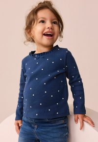 Marineblauwe lange mouw top met witte stippen, voorzien van een ruches halslijn en een klein strikdetail. Draagt met blauwe jeans.