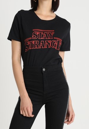 T-shirt en coton noir avec texte rouge "STAY STRANGE", manches courtes, col rond, associé à un jean noir taille haute avec des bandes latérales.