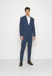 Navy blue suit jacket with two front pockets and a single button closure, paired with matching tapered trousers and black dress shoes.