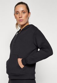 Femme à la peau claire et aux cheveux attachés, portant un sweat à capuche noir zippé avec des poches, debout devant un fond blanc uni.