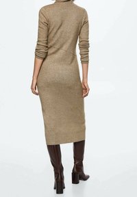 Vestido de punto en beige con cuello alto, mangas largas y un corte ajustado. La longitud llega hasta la mitad de la pantorrilla, combinado con botas marrones hasta la rodilla.