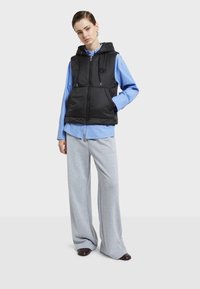 Musta pehmendatud vest kapuutsiga, millel on lukk, nöörid ja logo. Kanda heleda sinise särgi peal, koos hallide laiade pükstega.