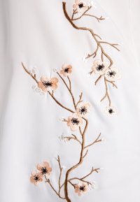 La tela blanca presenta un detallado bordado floral en suave rosa y negro, con ramas marrones y delicados acentos en blanco.