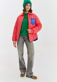 Žena v zelené čepici, zeleném svetru, korálově prošívané bundě s modrým kapsou, šedých džínách a botách se vzorem leopardí kůže stojí na bílém pozadí.