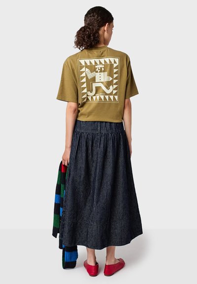 Donna che indossa una t-shirt grafica verde oliva e una lunga gonna a righe nere, tiene in mano una sciarpa a righe colorate, in piedi con le spalle rivolte verso la macchina fotografica.