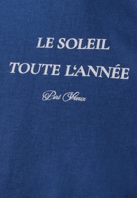 Modrá bavlněná tričko s bílým potiskem, na kterém je napsáno "LE SOLEIL TOUTE L'ANNÉE" a "Port Grieu" v kurzívě.