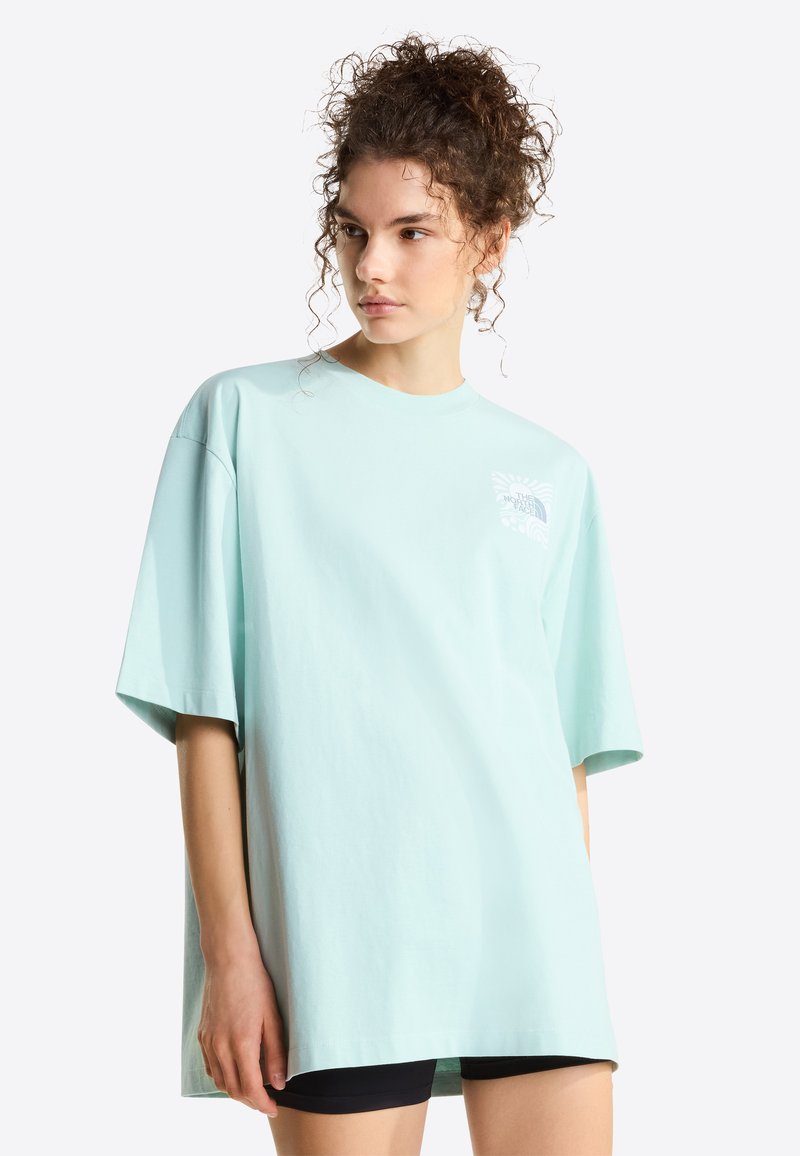 Frau mit lockigem Haar trägt ein übergroßes hellblaues T-Shirt mit kurzen Ärmeln und schwarze Shorts, schaut seitlich vor einem schlichten Hintergrund.