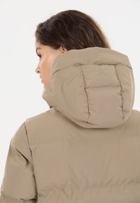 Beige pufferjakke med en høj, struktureret hætte. Stoffet ser blødt ud med en glat tekstur, der har diskrete syninger og et logodetalje på ryggen.