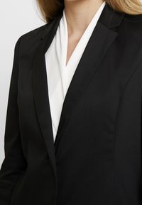 Americana negra con un corte entallado, que presenta un forro interior blanco y solapas de pico. Textura suave y diseño clásico.