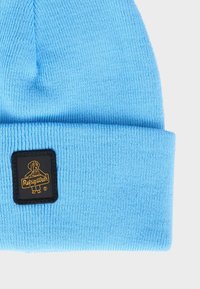 Berretto a maglia azzurro chiaro con risvolto, caratterizzato da una toppa quadrata nera con una grafica di un cane e la scritta "RefijigWear" in lettere dorate.