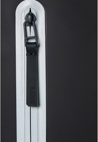 Tirette de fermeture éclair noire avec le logo « RAINS » embossé, fixée à une fermeture éclair blanche sur un fond en tissu noir.