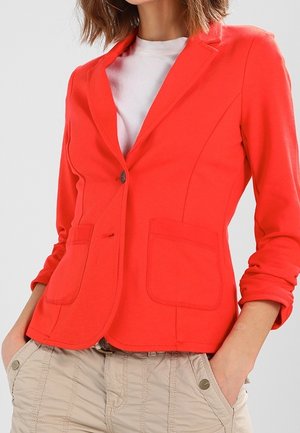 Femme portant un blazer rouge vif sur une chemise blanche avec un pantalon beige, les mains dans les poches et les manches du blazer légèrement retroussées.