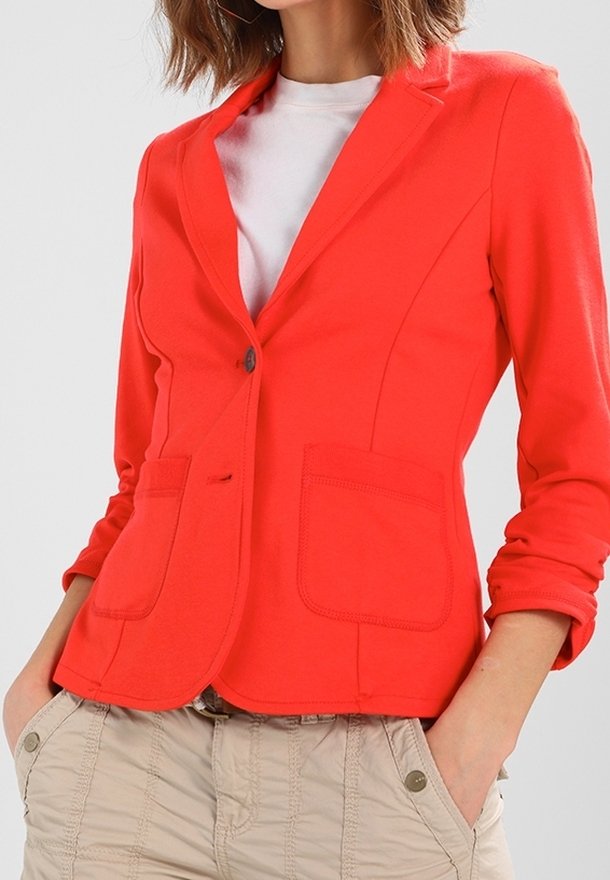 Femme portant un blazer rouge vif sur une chemise blanche avec un pantalon beige, les mains dans les poches et les manches du blazer légèrement retroussées.