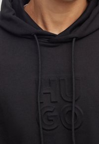 Felpa nera con cappuccio realizzata in un morbido tessuto, caratterizzata da un logo "HUGO" in rilievo sul davanti. Include cordini e un colletto classico.