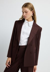 Mujer con blazer y pantalones de rayas marrones, de doble botonadura, camisa blanca de botones, mirando hacia abajo, con grandes pendientes redondos, sobre fondo blanco.
