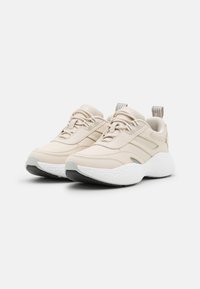 Pár béžových tenisek Calvin Klein s tlustými bílými texturovanými podpatky a vícerozvlekovými poutky, nakloněné doprava na bílém pozadí.