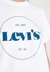 Bílý bavlněný tričko s velkým modrým logem Levi's uvnitř kruhu, s nápisem "EST. 1873" a "SF / CA." vytištěným nad a pod logem.
