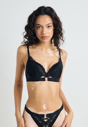 Black lace bra with molded cups, gold accent hardware, and front closure. Matching lace thong with strap details completes the set.