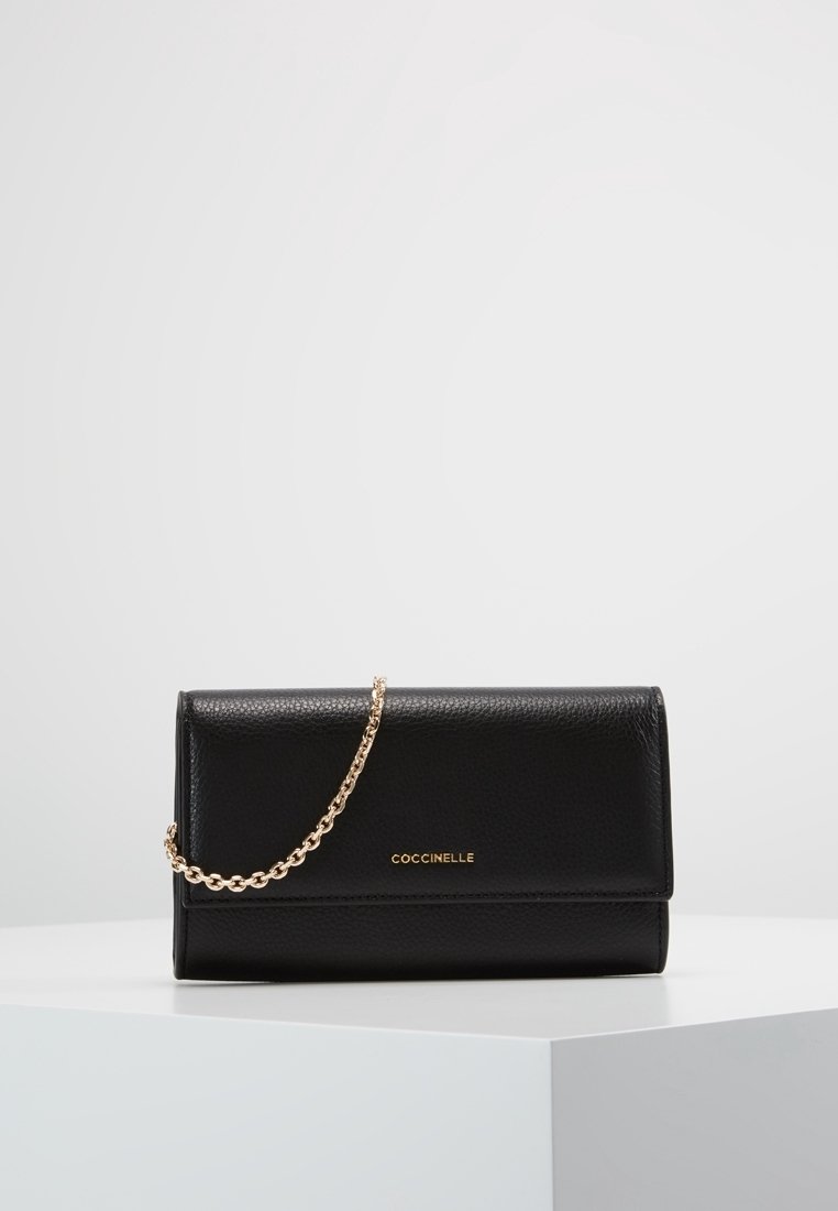 Pochette en cuir noir avec une bandoulière chaîne dorée et le logo "COCCINELLE" doré sur le devant, exposée sur une surface blanche avec un fond uni.