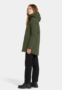 Vrouw in een olijfgroene hoodiejack, zwarte broek en zwarte laarzen, staand in profiel tegen een neutrale achtergrond.