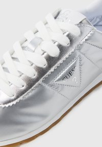 Baskets synthétiques argentées avec lacets blancs, dotées d'un écusson logo triangulaire et d'une semelle texturée. Finition lisse et design épuré.