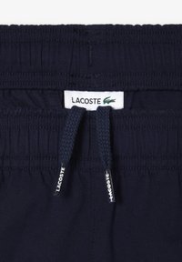 Pantaloni corti con elastico in vita di colore blu navy, dotati di cordini di regolazione, con etichetta bianca Lacoste e tessuto testurizzato. Nessuna tasca visibile.