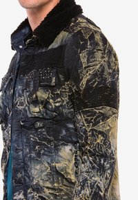 Schwarze und gelbe, zerknitterte Stoffjacke mit einem Kunstfellkragen, Fronttaschen und genoppten Akzenten an den Schultern. Texturiertes, robustes Design.