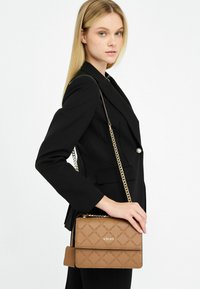 Braune synthetische Leder Tasche mit Diamantmuster, goldenen Beschlägen und einer Kettenriemen. Die Tasche verfügt über einen Klappenverschluss und ein geprägtes Logo.